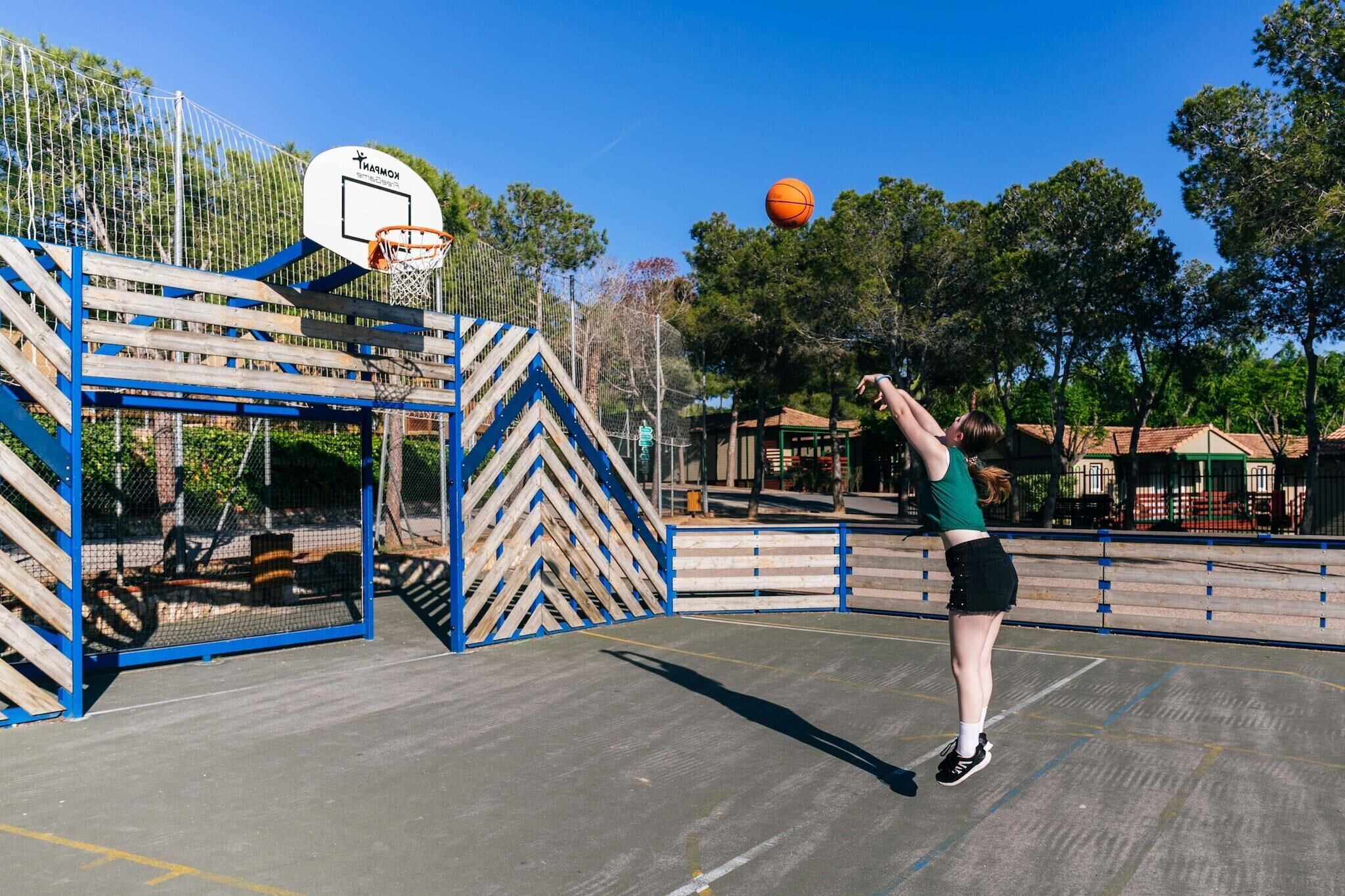 Un terrain de basket avec un panier, une clôture en bois et des arbres en arrière-plan.
