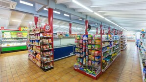 A supermarket aisle with shelves stocked with various products, refrigerated sections, and a visible checkout area.