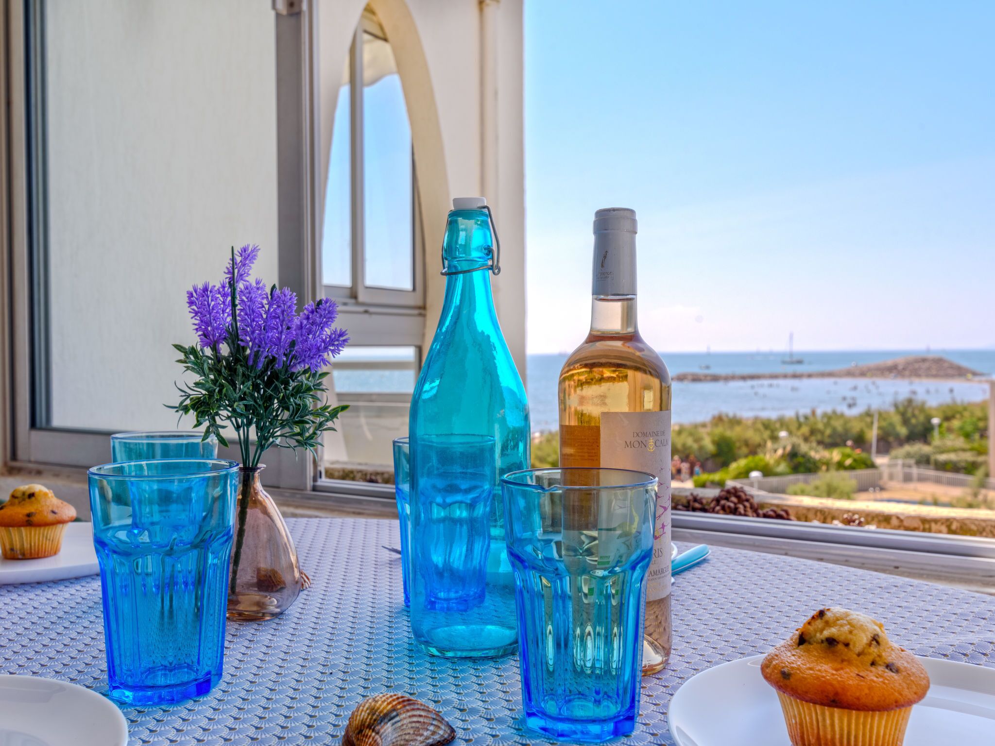 Een tafel met een fles Domaine de Mon Calme, glazen, een muffin en een decoratieve vaas met paarse bloemen.