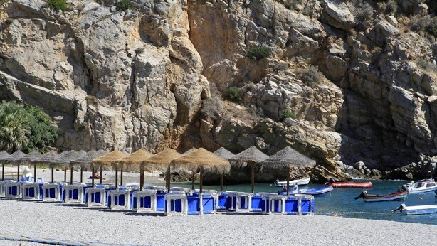 En strand med rækker af blå strandstole og stråparasoller, klippestykker i baggrunden og både i vandet.