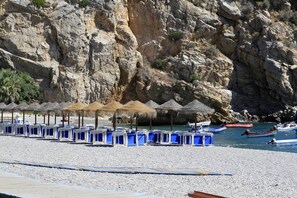 A beach with rows of blue beach chairs and thatched umbrellas, rocky cliffs in the background, and boats in the water.