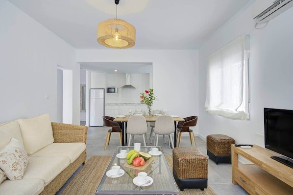 A modern living room with a sofa, coffee table, TV stand, and a dining area with chairs and a table.
