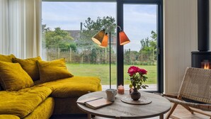 Ein gemütliches Wohnzimmer mit einem gelben Sofa, einem runden Holztisch und einem Korbstuhl. Im Hintergrund ist ein Kamin zu sehen.