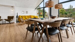 Ein moderner Essbereich mit einem großen Holztisch und Stühlen, einem gelben Sofa und großen Fenstern mit Blick ins Freie.