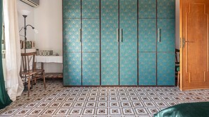 A room with a patterned floor, a large wardrobe with multiple doors, a wooden door, a chair, and a table with a lamp.