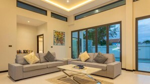 Ein modernes Wohnzimmer mit einem grauen Ecksofa, einem Glastisch und großen Fenstern mit Blick ins Freie.