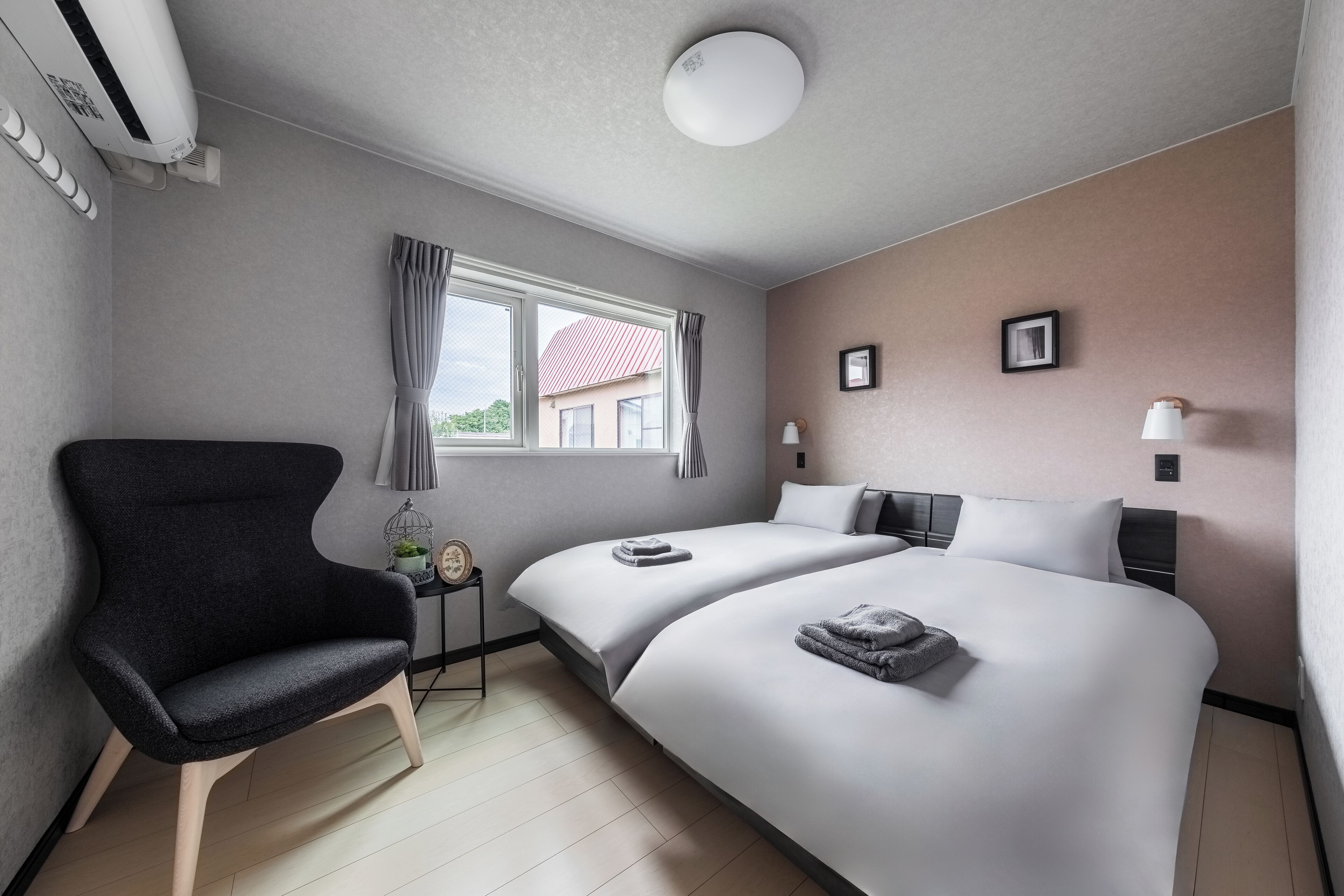 Habitación de hotel con dos camas, una silla, una ventana con cortinas, una lámpara en el techo y aire acondicionado instalado en la pared.