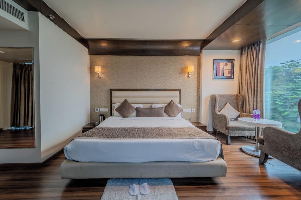 Une chambre d’hôtel moderne, dotée d’un grand lit, d’une chaise, d’une petite table et offrant une vue sur la verdure par la fenêtre.