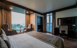 A hotel room with a bed, two armchairs, a small table with bottles, a TV, and a large window with curtains. - Hotel Aditya Park Regent (Jaipur)