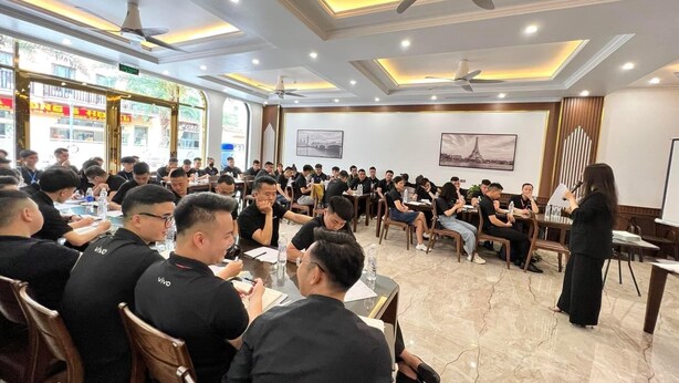 A conference room with attendees seated at tables, a presenter at the front, and a large window with a view of the street. - Quynh Anh Hotel Ha Long (Bai Chay)