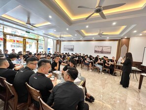 Un salón de conferencias con asistentes sentados en mesas, un presentador al frente y un amplio ventanal con vistas a la calle.