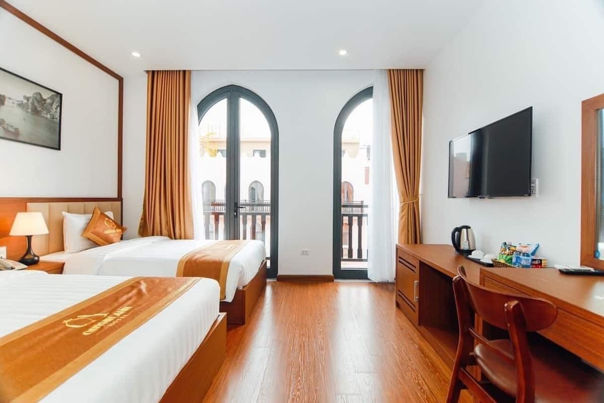 A hotel room with two beds, a desk, a TV, and a balcony with curtains.