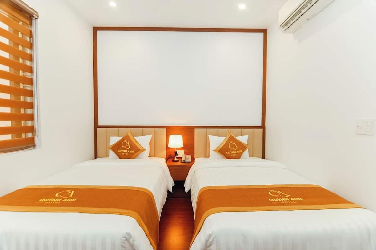 A hotel room with two beds, a nightstand with a lamp, and a large window with blinds.