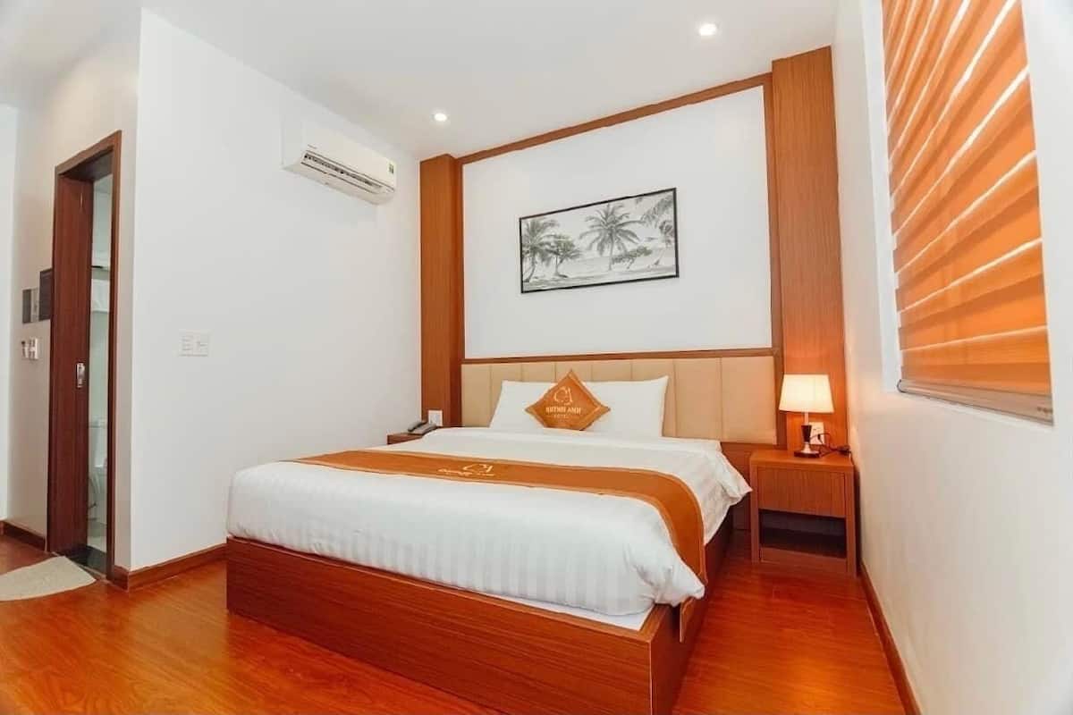 A hotel room with a wooden bed, a nightstand with a lamp, a framed picture on the wall, and an air conditioning unit.