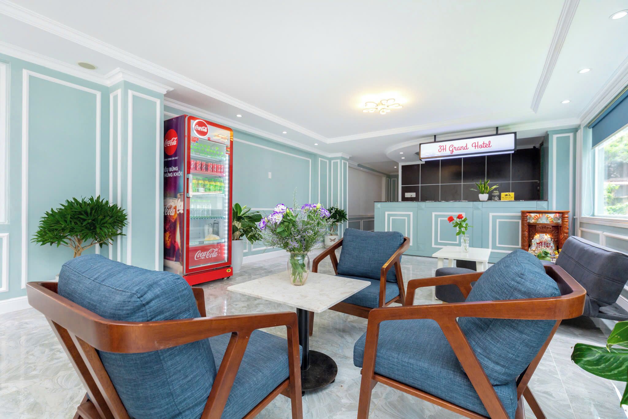 Un hall de hotel con una máquina expendedora de Coca-Cola, un mostrador de recepción y un área de descanso con sillones tapizados en azul.