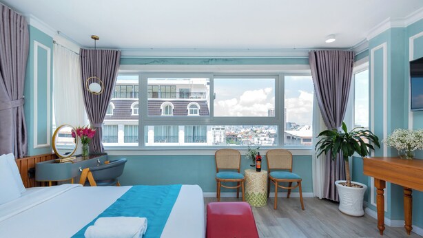 A hotel room with a bed, a desk, two chairs, a small table, a potted plant, and a view of the city.