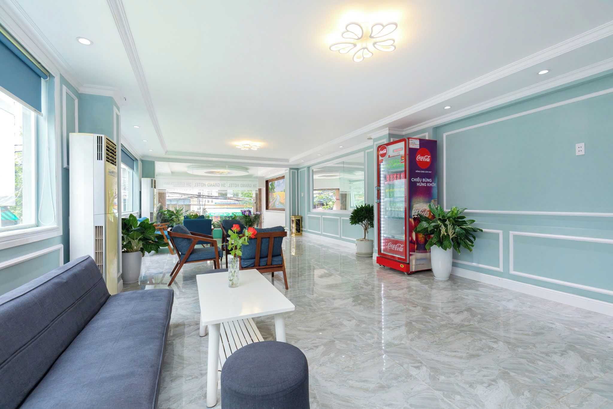 A hotel lobby with a sofa, chairs, a table, potted plants, and a Coca-Cola vending machine.