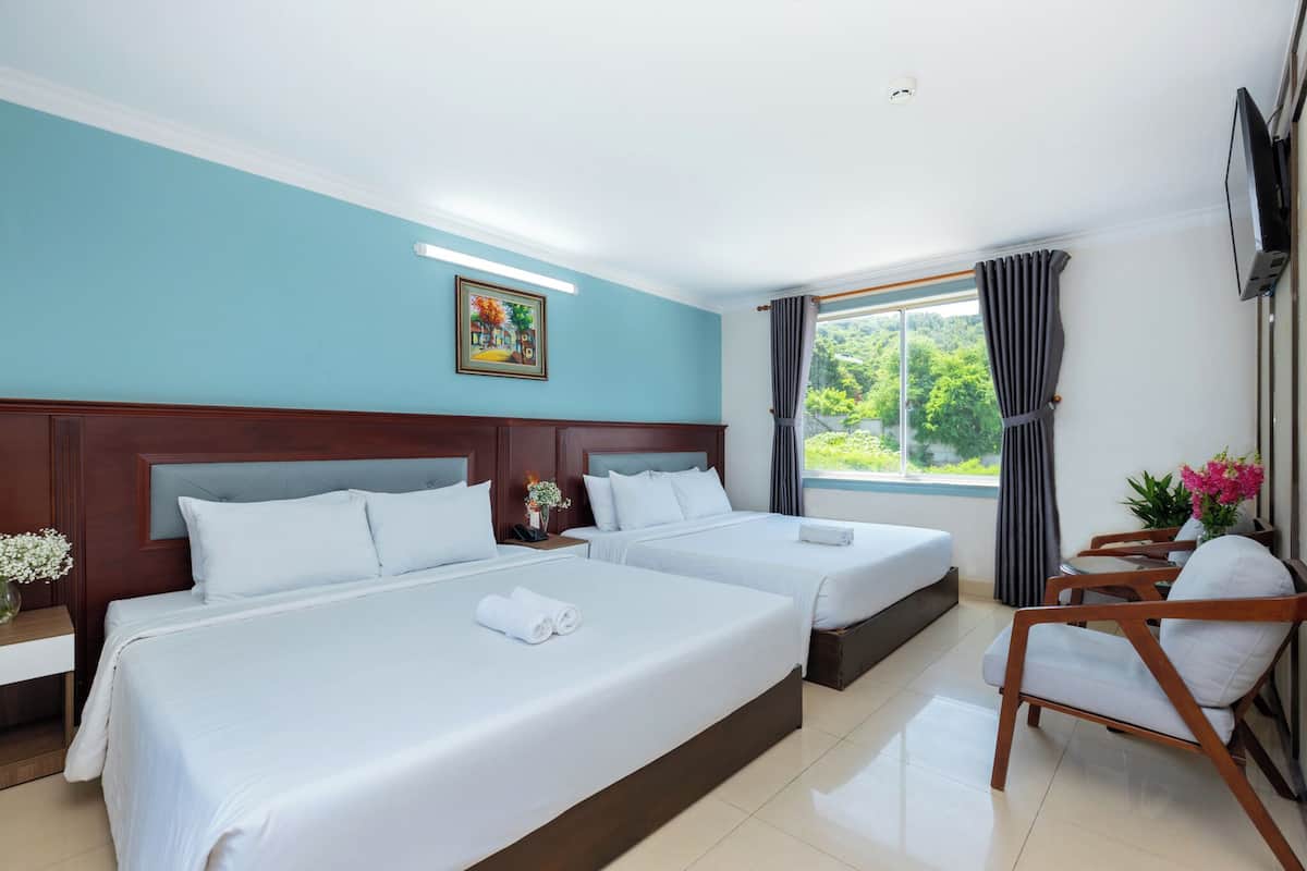A hotel room with two beds, a wooden headboard, a TV, a window with curtains, and a view of greenery outside.