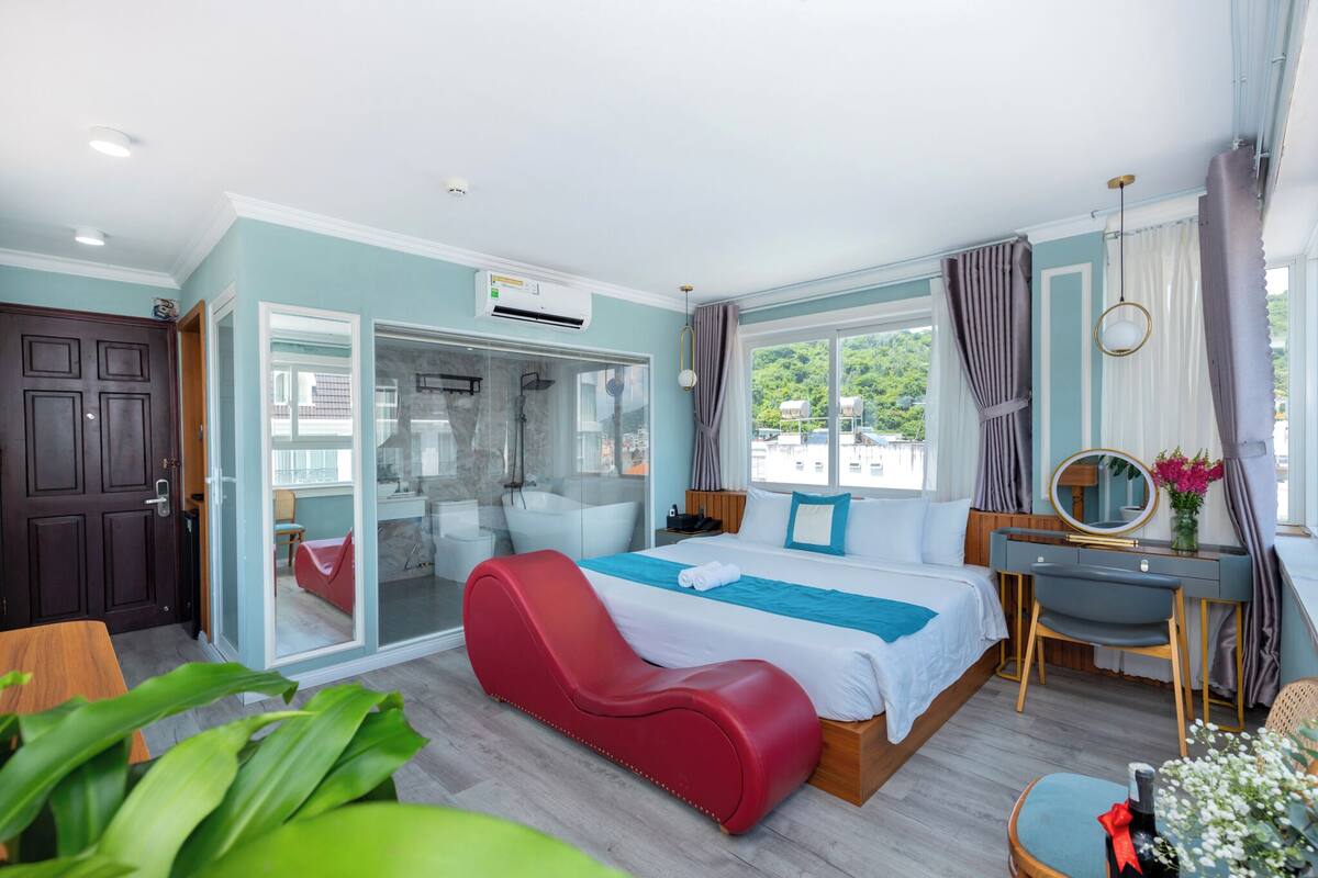 A modern bedroom with a bed, a red lounge chair, a desk, and a glass shower area.