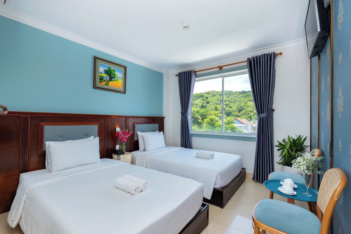 A hotel room with two beds, a small table with a chair, a window with curtains, and a painting on the wall.
