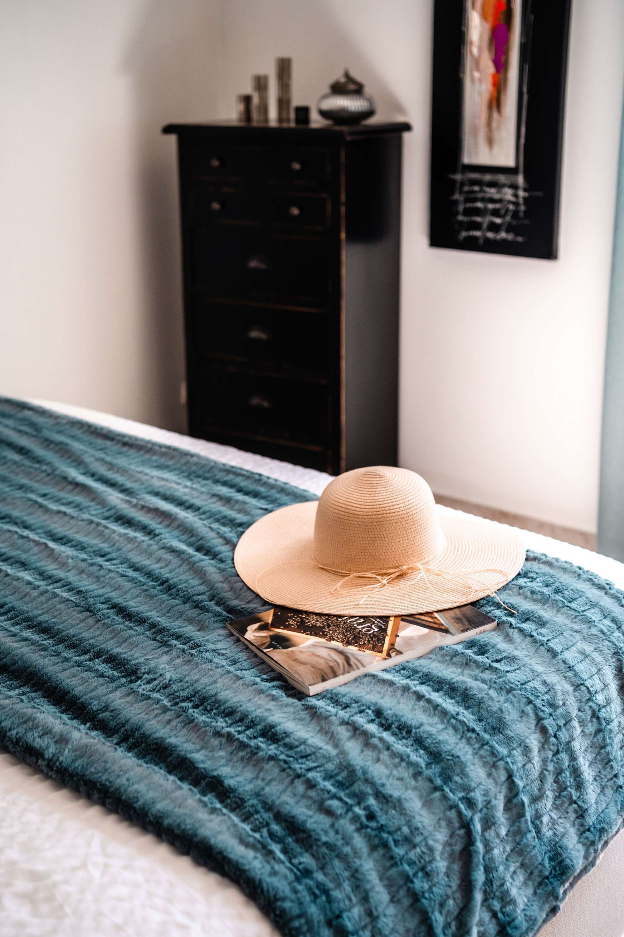 Una cama con una manta verde azulada, un sombrero de paja y una revista encima.