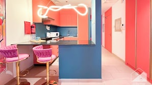 A modern kitchen with a central island, pink bar stools, and a neon light fixture. - Colorful Studio With Balcony & City Views (Dubai)
