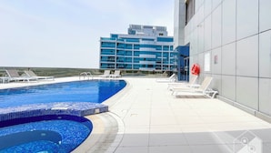 A rooftop swimming pool with lounge chairs and a modern building in the background. - Colorful Studio With Balcony & City Views (Dubai)