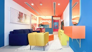 A modern living room with a blue sofa, yellow armchairs, and a TV. There is a kitchen area in the background with a red wall and a mirror. - Colorful Studio With Balcony & City Views (Dubai)