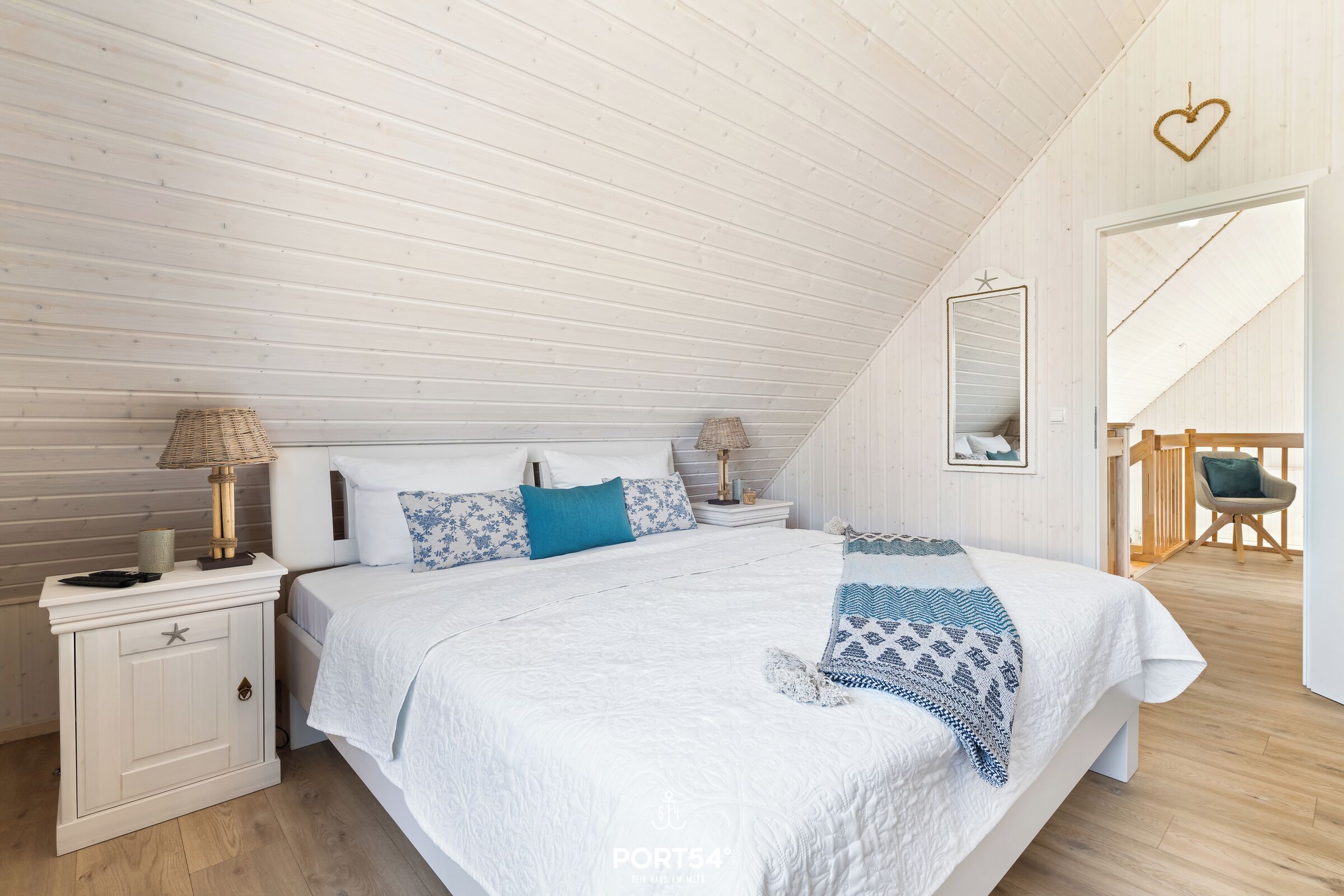 A bedroom with a sloped ceiling, a bed with white bedding, two bedside tables with lamps, a mirror, and a chair.