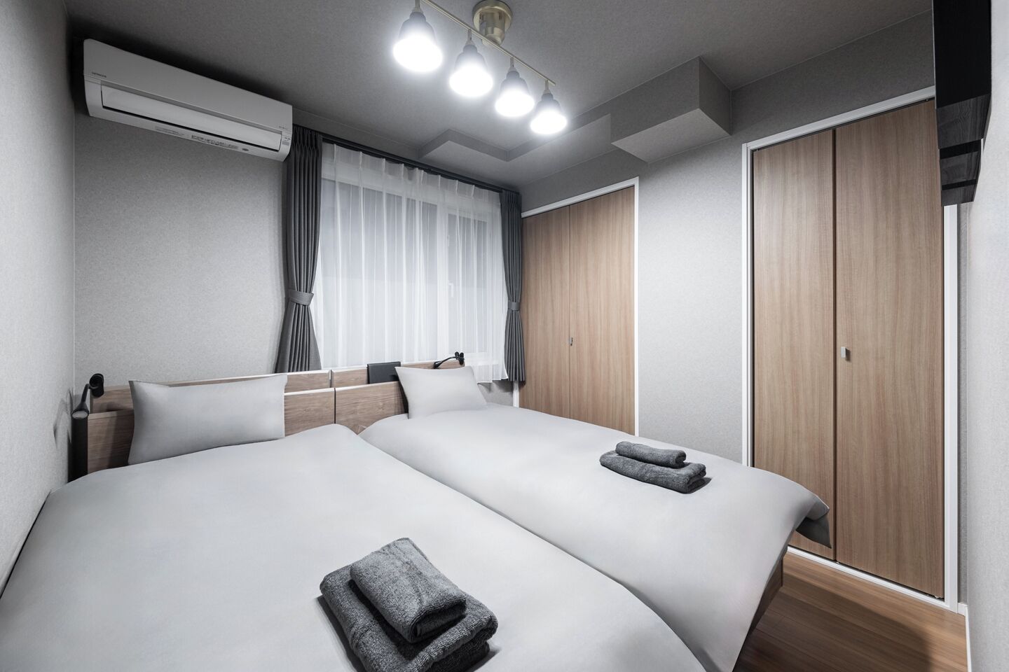 A modern hotel room with two beds, a wooden wardrobe, and an air conditioning unit.