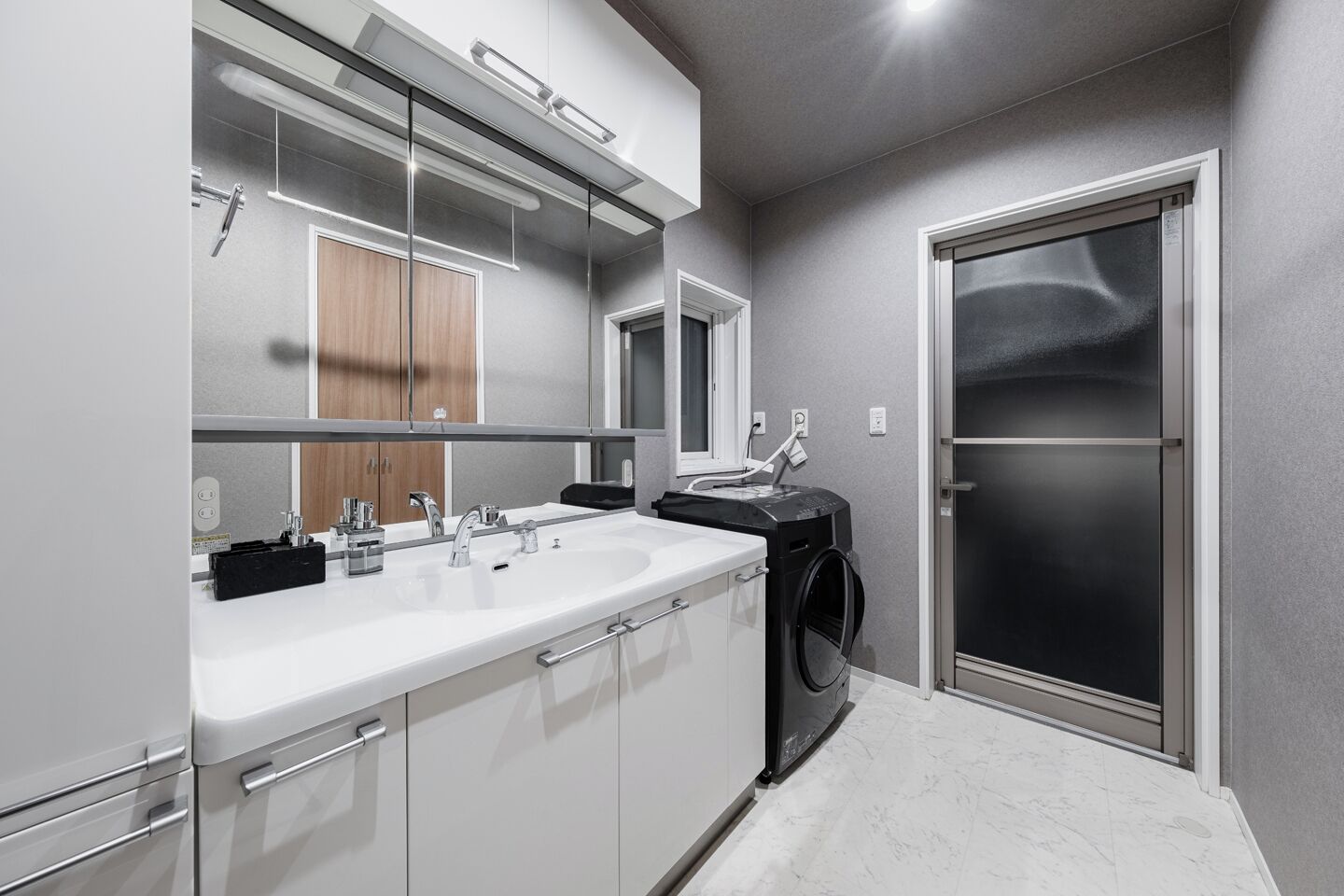 A modern bathroom with a large mirror, two sinks, a black washing machine, and a frosted glass door.