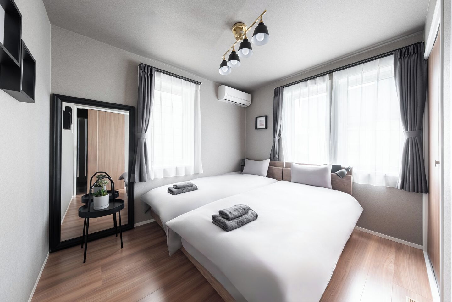 Une chambre d’hôtel moderne avec deux lits, une petite table agrémentée d’une plante, un luminaire au plafond et de grandes fenêtres avec des rideaux.