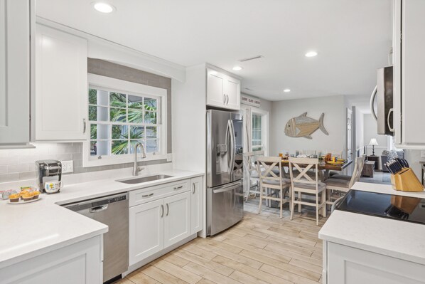 A modern kitchen with white cabinetry, a stainless steel refrigerator, a dishwasher, and a dining area with a wooden table and chairs. - 10 Willow Oak @ Sea Pines| Ocean Course View | Pool | Beach Club (Hilton Head Island)