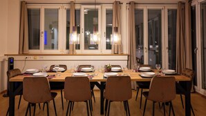 A dining area with a long wooden table set for six, surrounded by six chairs, with a city view through large windows.