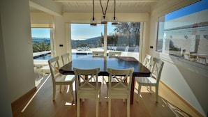 A dining area with a large table and chairs, a hanging light fixture, and a view of a pool and mountains through large windows. - Spacious 4 Bedroom Villa & Apartment Suite, Private Pool and Fantastic Views (Calonge)