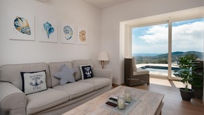 A living room with a sofa, a coffee table, and a view of a swimming pool and mountains through large windows. - Spacious 4 Bedroom Villa & Apartment Suite, Private Pool and Fantastic Views (Calonge)