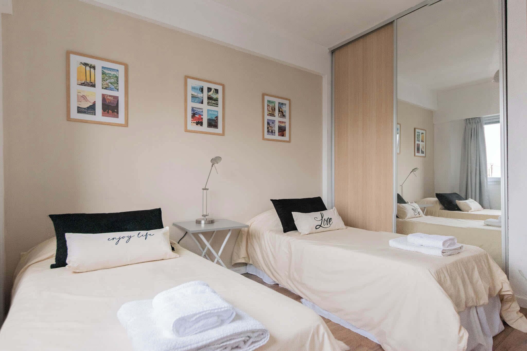 A hotel room with two beds, each with a pillow and a towel. There are framed pictures on the wall and a mirror reflecting the room.