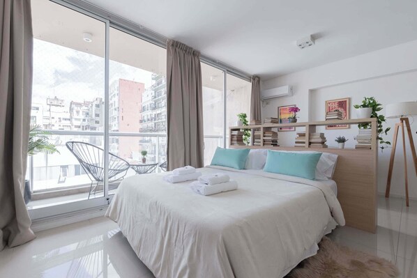 Un dormitorio moderno con un ventanal grande, una cama con ropa de cama blanca, un cabecero de madera y una mesita de noche con una lámpara.