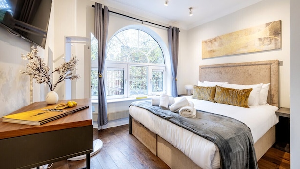 A bedroom with a large bed, a desk with a vase of flowers, and a window with curtains. - Prime 3 Bedrooms Flat – Perfect for Families & Groups (Greater London)