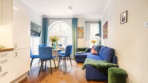 A modern living room with a blue sofa, a dining table with chairs, and a kitchen area with white cabinets. - Prime 3 Bedrooms Flat – Perfect for Families & Groups (Greater London)