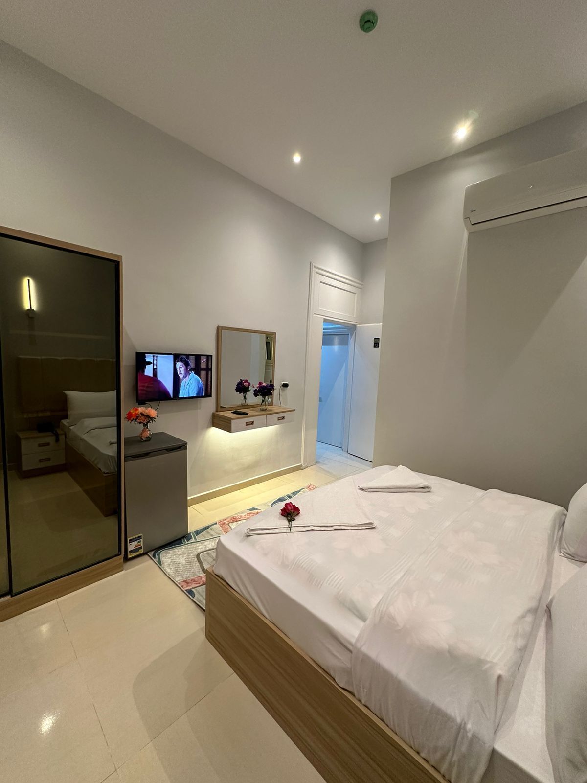 A modern hotel room with a large bed, a TV, a mirror, a wardrobe, and a small desk with a vase of flowers.