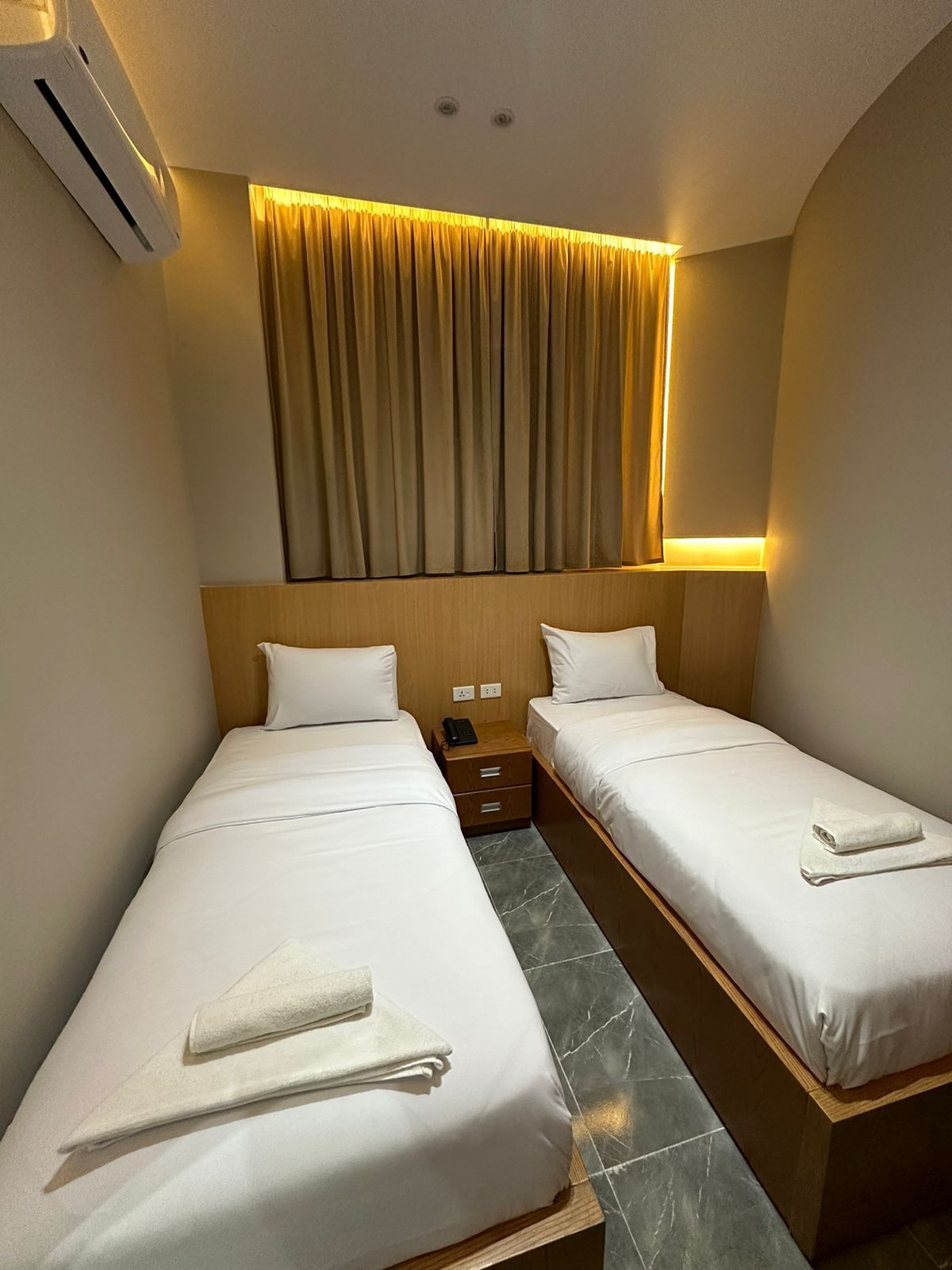 A hotel room with two single beds, a bedside table with a phone, and a curtain above the beds.