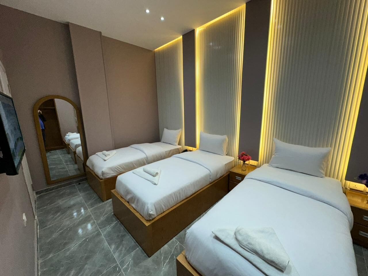 Une chambre d’hôtel avec trois lits, un grand miroir et une petite table sur laquelle est posé un vase de fleurs.
