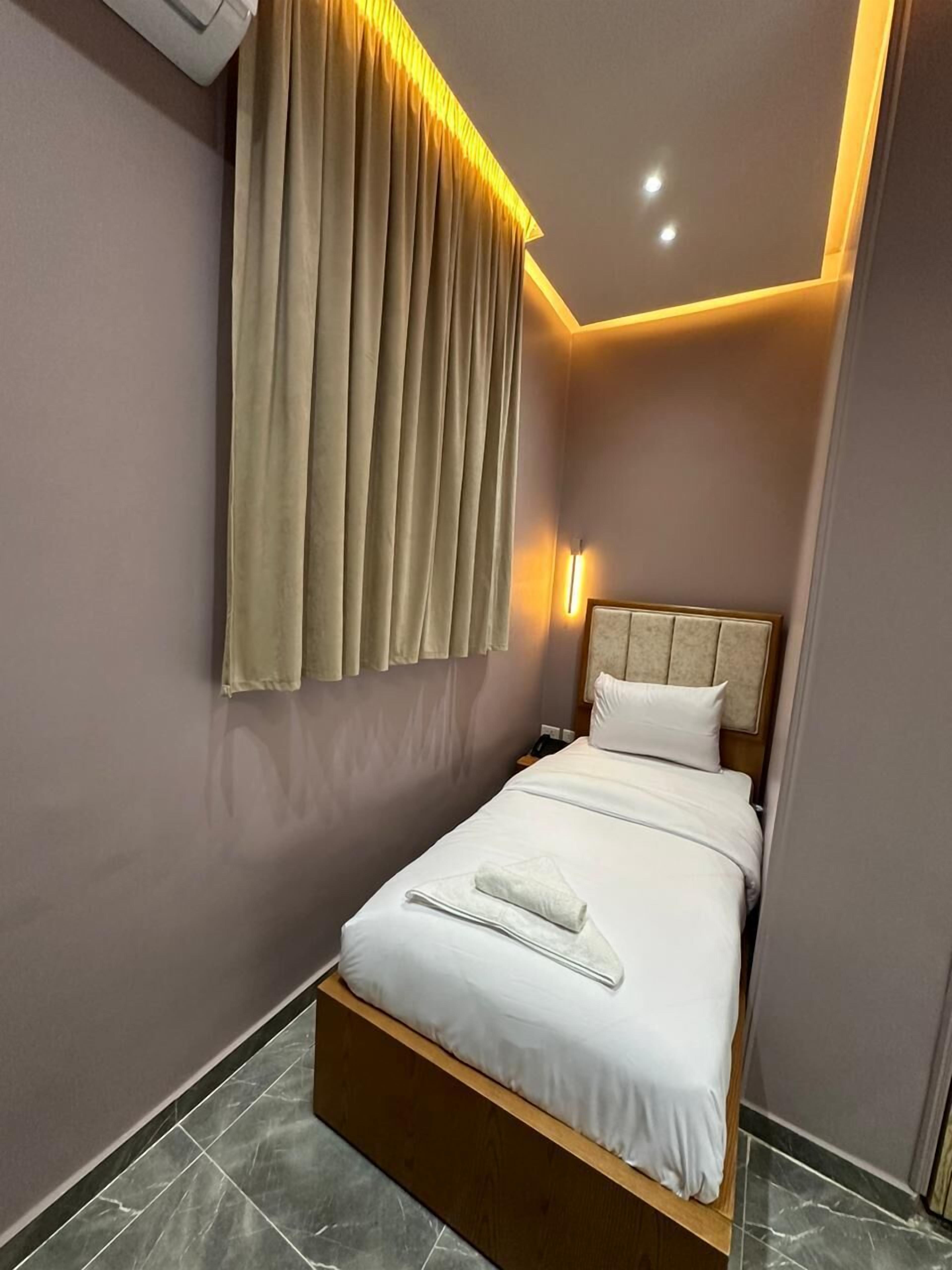A neatly made bed with white linens and a folded towel, a headboard, and a wall-mounted light fixture.