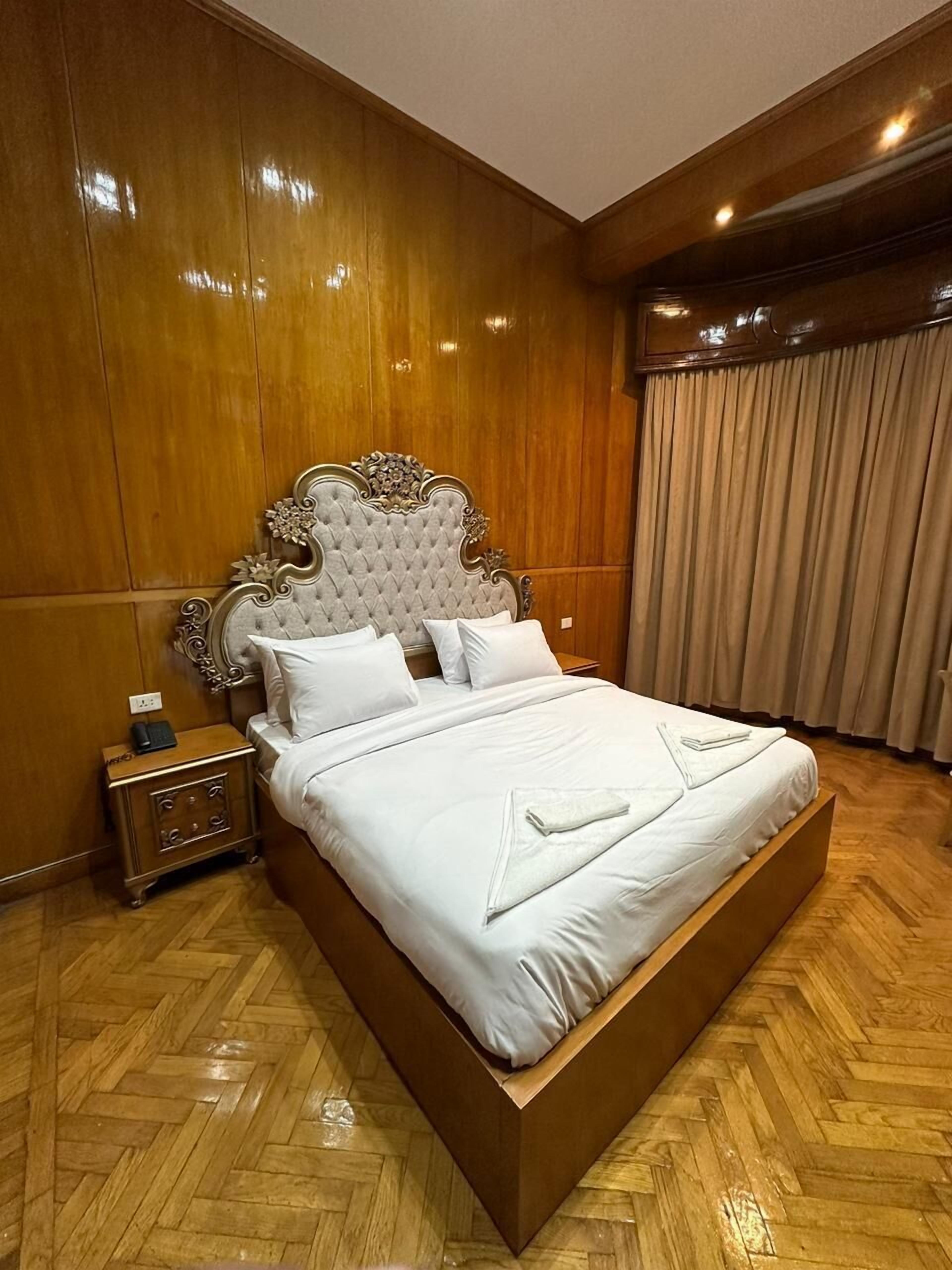 A large, ornate bed with a tufted headboard, a wooden nightstand, and a herringbone-patterned wooden floor.