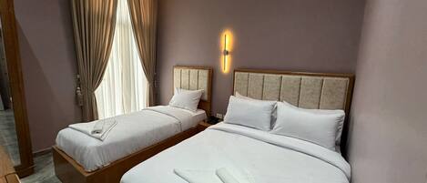 A hotel room with two beds, a large window with curtains, a wall-mounted light, and a wooden bench.