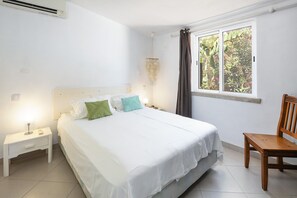 A bedroom with a bed, a nightstand, a chair, a window with curtains, and an air conditioning unit.