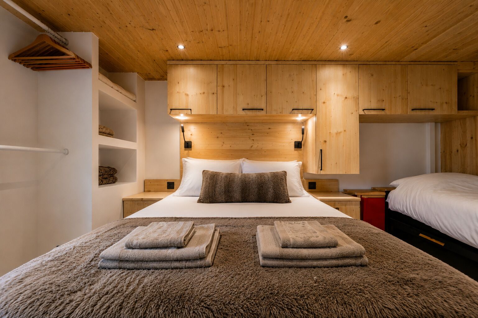 Een slaapkamer met een houten plafond, een bed met wit beddengoed, een hoofdbord met ingebouwde verlichting en een nachtkastje in het rood.