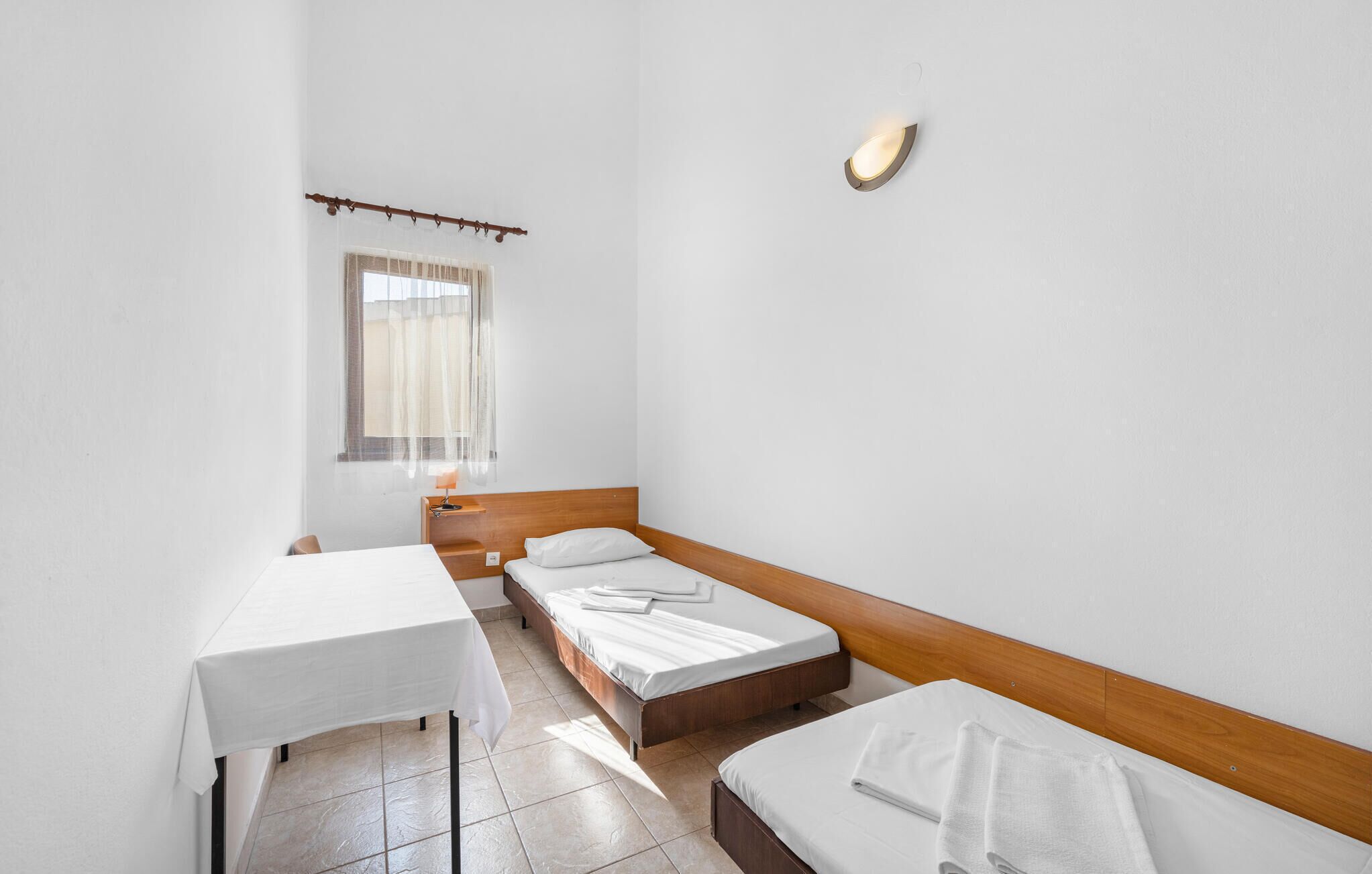 Une petite chambre propre, avec deux lits simples, une table et une fenêtre ornée de rideaux légers.
