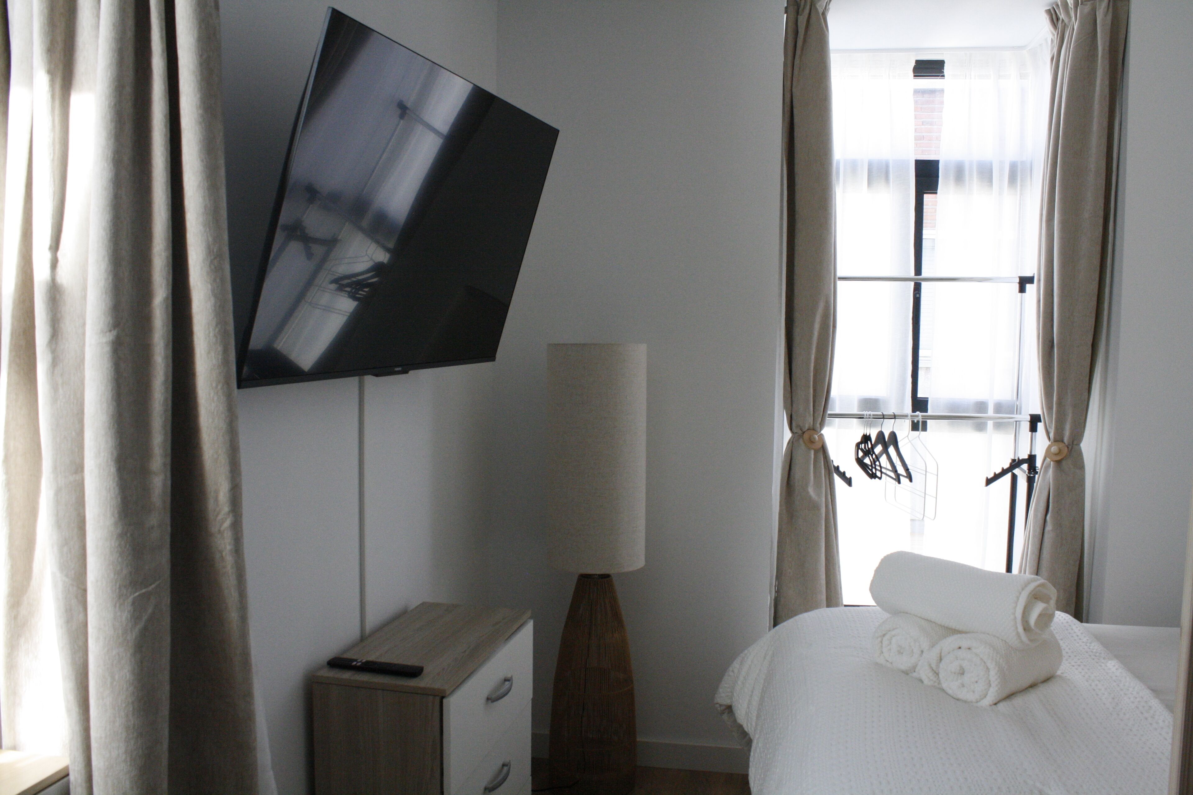 A hotel room with a flat-screen TV mounted on the wall, a bedside lamp, a neatly made bed with rolled towels, and a window with curtains.